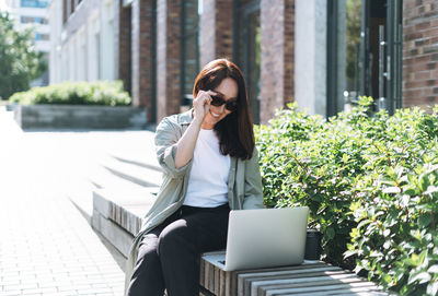 Adult woman forty years in stylish shirt working on laptop at public place on bench at city street