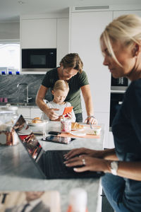 Woman using laptop while man and son holding smart phone in kitchen at home