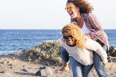 Cheerful woman carrying friend on back against sea