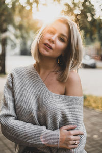 Portrait of young woman with head cocked standing on sidewalk in city during sunset