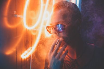 Young man wearing sunglasses and smoking while standing by illuminated lights at night