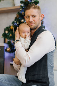 A happy father holds a newborn baby in his arms against the background of a christmas tree 