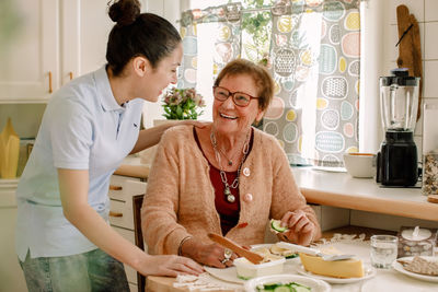 Cheerful senior woman talking with female caregiver in kitchen at nursing home