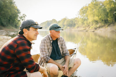 Male friends looking away while sitting on boat at lakeshore