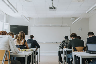 Rear view of students studying in classroom with blank whiteboard