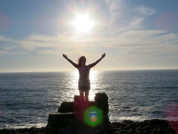 Woman standing with arms outstretched on rocks at beach against sky on sunny day