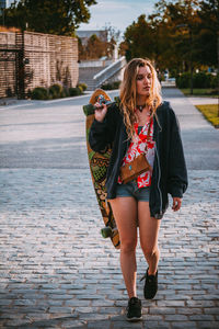 Young woman holding skateboard looking away walking on street in city