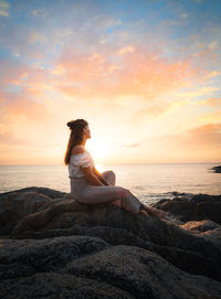 Man sitting on rock by sea against sky during sunset