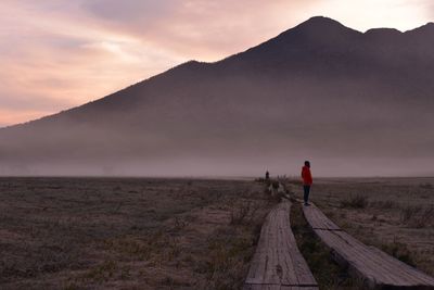 Person standing on wooden pathway against mountains during sunset