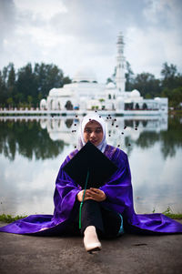 Portrait of woman holding mortarboard while sitting against lake with mosque in background