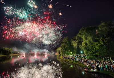 Crowd looking at firework display over river against sky at night