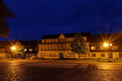 Illuminated street by buildings against sky at night