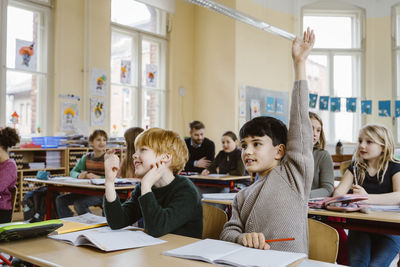 Boy with hand raised answering during lecture while sitting by male friend in classroom