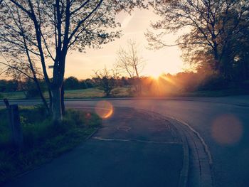 Road by bare trees against sky during sunset