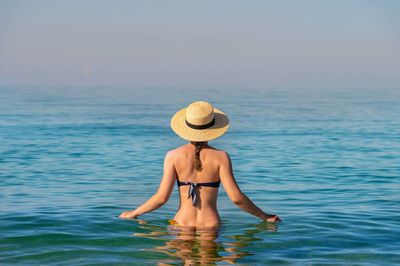 Back view of a slender caucasian young woman standing waist-deep in calm transparent sea water in a