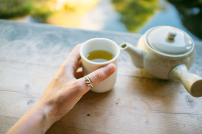 Cropped hand holding green tea cup by teapot on table