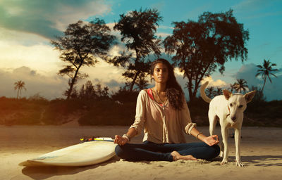 Portrait of young woman with dog meditating while sitting at beach against sky during sunset