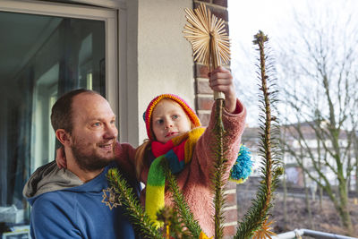 Smiling man carrying daughter wearing knit hat decorating christmas tree