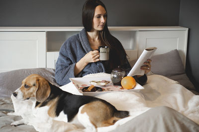 Dog beagle and smiling woman female eat breakfast and reading book in the bedroom of the house