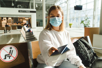 Young woman traveler in medical mask sits in the waiting room and holds out passport and ticket