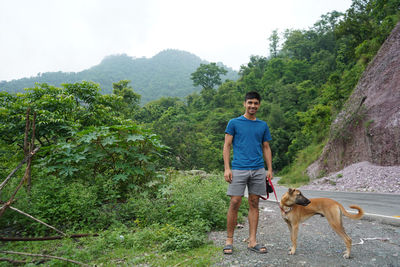 Young man with his dog standing beside a road with blurred mountains in the background.