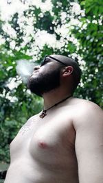 Low angle view of shirtless man smoking while standing against trees