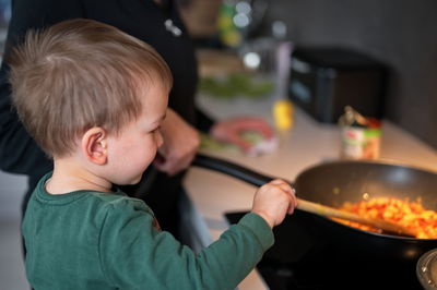 Cute little toddler at the stove cooking vegetables with his mother