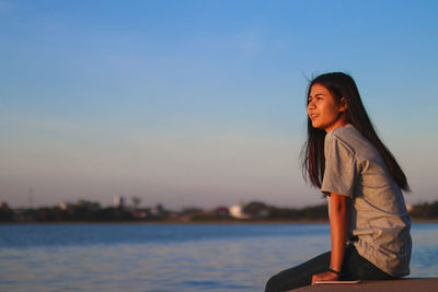 Side view of smiling young woman sitting on retaining wall by sea against clear sky during sunset
