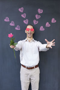 Man with heart shape paper on eyes standing against blackboard