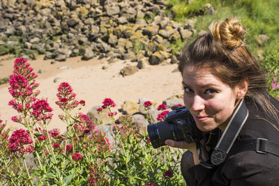 Portrait of young woman holding camera by flowering plants on field