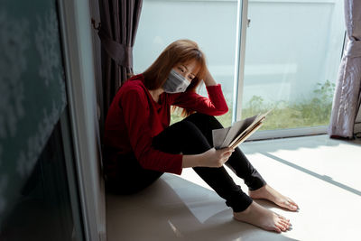 Young woman using mobile phone while sitting on window