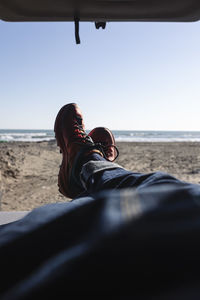 Man's legs in blue jeans and hiking shoes lying inside a van on the beach.