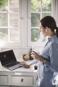 Side view of female professional using laptop on counter while drinking water at home office