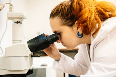 Side view of concentrated female scientist in white medical uniform looking through modern microscope while examining samples in light laboratory