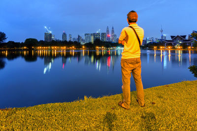 Rear view of man standing at riverbank against illuminated cityscape at night