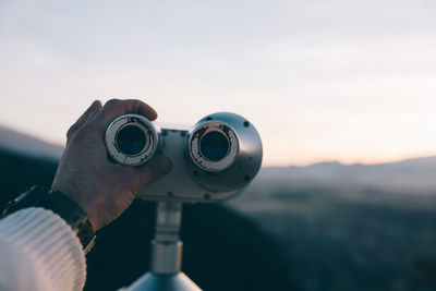 Close-up of hand holding binoculars against sky during sunset