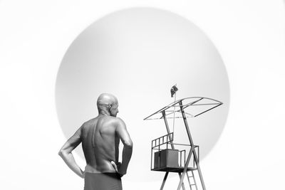 Rear view of shaved head man with hands on hip against lookout tower