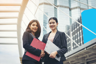 Portrait of smiling businesswomen with clipboards standing against buildings in city 