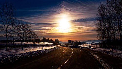 Road by silhouette bare trees against sky during sunset