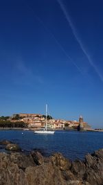 Sailboats moored on sea by buildings against blue sky