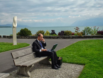 Full length of businessman using laptop while sitting on bench against sea