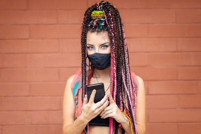 Portrait of woman holding smart phone while standing against brick wall