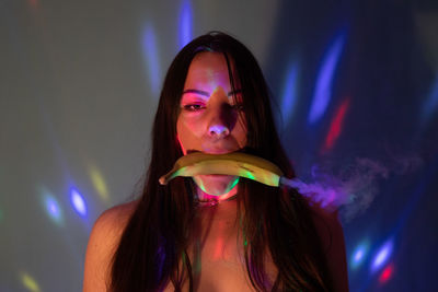 Portrait of young woman holding kiwi mouth against lights on wall