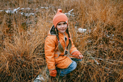 Happy girl in orange coat sits in forest among dry grass and golden trees, outdoor, fall vibes