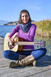 Portrait woman playing guitar while sitting on pier by lake