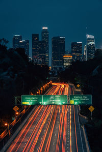 Light trails on road by illuminated buildings against sky at night