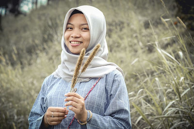 Portrait of smiling girl wearing hijab holding plant while standing on grassy field