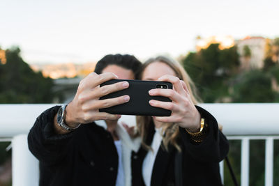 Joyful young multiracial couple in casual wear taking selfie on mobile phone while standing together on bridge with green trees and city buildings in background