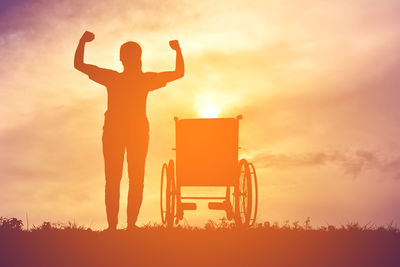 Silhouette woman flexing muscles while standing wheelchair on field during sunset