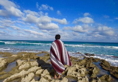 Rear view of man with blanket standing at rocky beach against sky